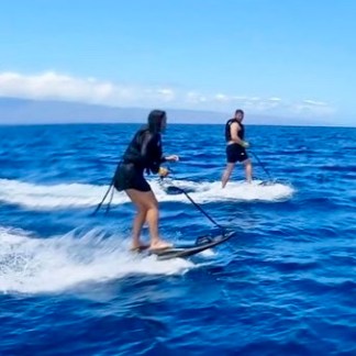 Two people on motorized surfboards riding waves in the ocean under a clear blue sky.