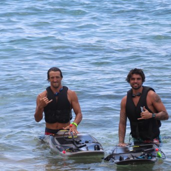 Two men in life vests holding jetboards in the ocean, gesturing a shaka sign with their hands.