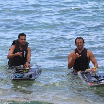 Two people in water holding jet boards, wearing life vests, making hand gestures.