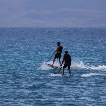 Two people stand-up paddleboarding on the sea near a sailboat.