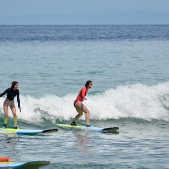 Two women surfing on small waves near the shore with a calm ocean in the background.